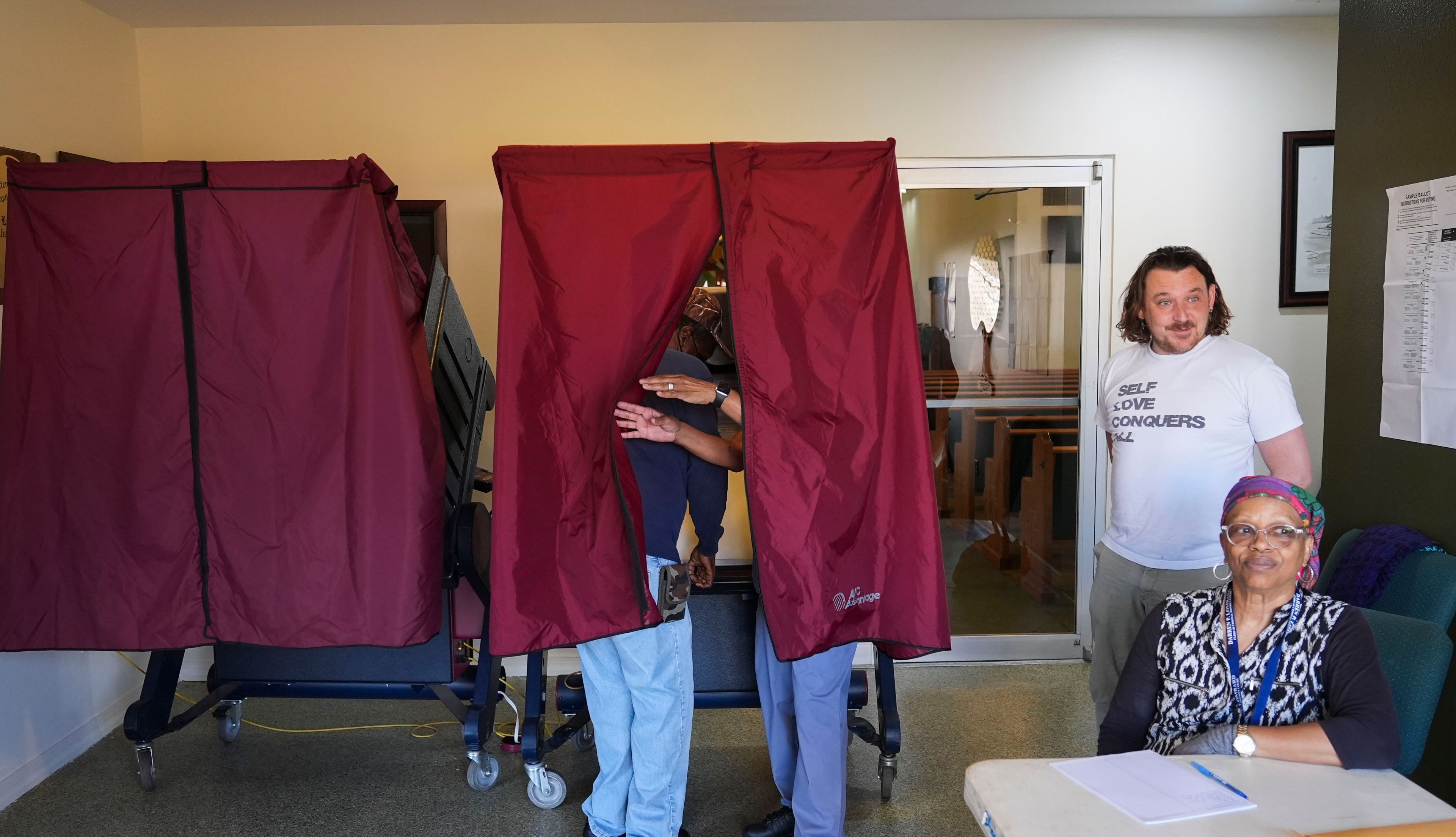 Precinct commissioners Ruby Augustine and Aloysius Cunningham, right, look on as Linda Matthews assists her husband Johnny Matthews, Sr. inside a voting booth as they vote at the Greater Evergreen Baptist Church in New Orleans on Election Day, Tuesday, Nov. 5, 2024. (AP Photo/Gerald Herbert)