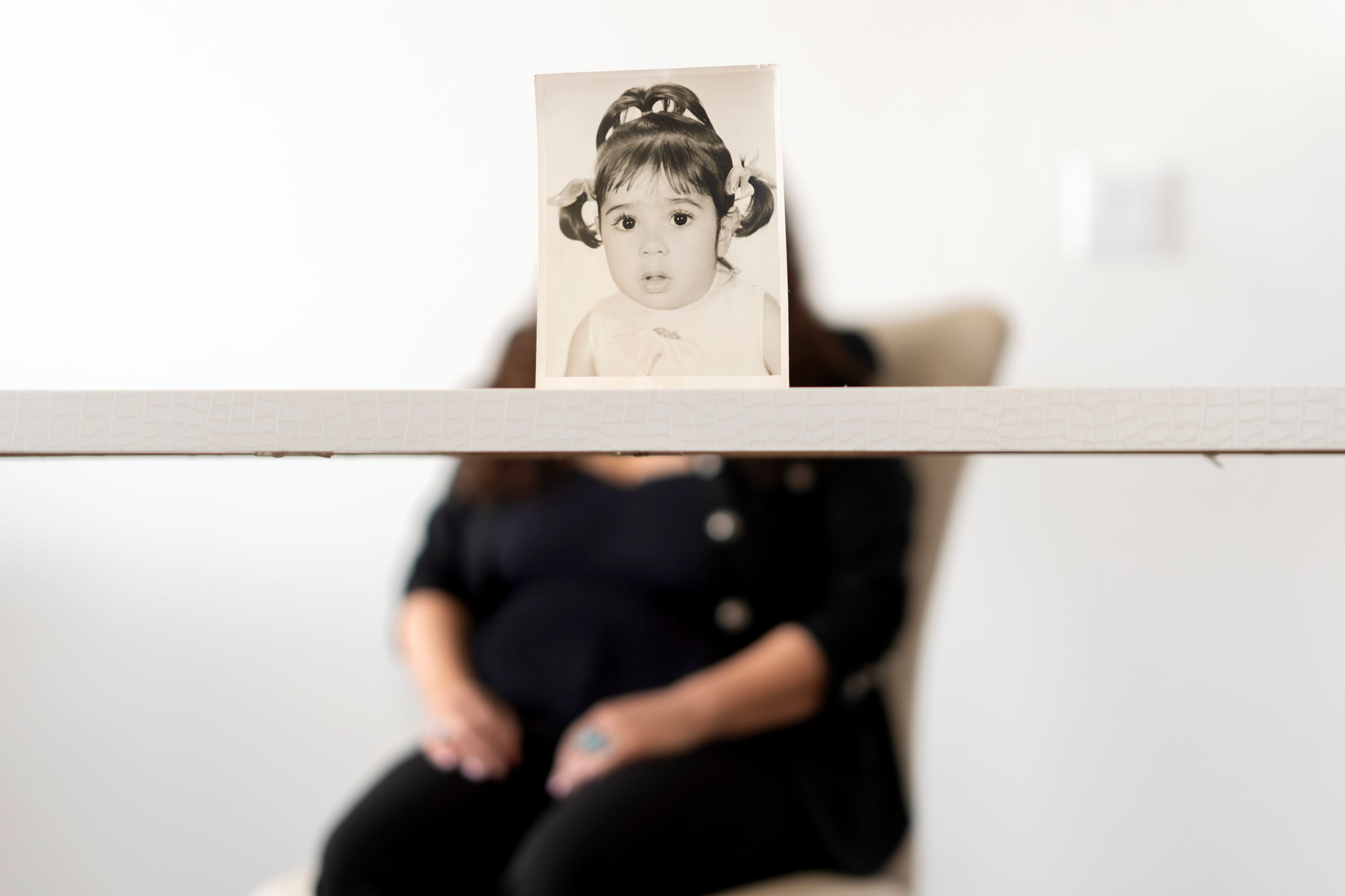 Buttons, who the Associated Press is referring to only by her childhood nickname because of her legal status, sits for a portrait behind her baby photo taken before she was adopted from Iran to a family in America, Monday, June 24, 2024, in Henderson, Nev. Buttons is one of thousands of children adopted from abroad by American parents, many of them military service members, who were left without citizenship by loopholes in American law that Congress has been aware of for decades, yet remains unwilling to fix. (AP Photo/David Goldman)