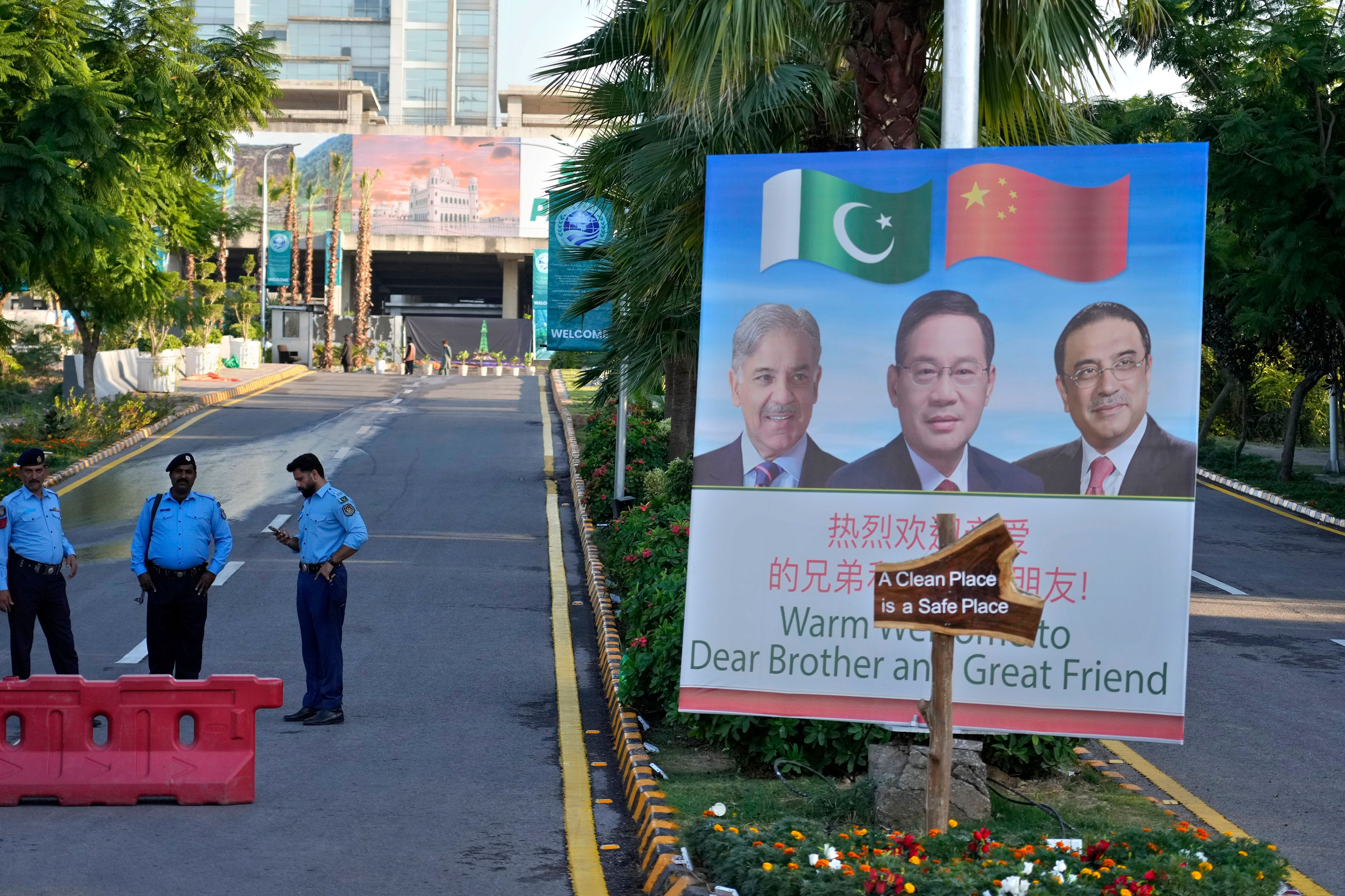 Police officers stand guard next to a welcoming billboard with portraits of China's Premier Li Qiang, center, Pakistan's Prime Minister Shehbaz Sharif and President Asif Ali Zardari, displayed at a road leading to the venue of the upcoming Shanghai Cooperation Organization (SCO) summit in Islamabad, Pakistan, Sunday, Oct. 13, 2024. (AP Photo/Anjum Naveed)