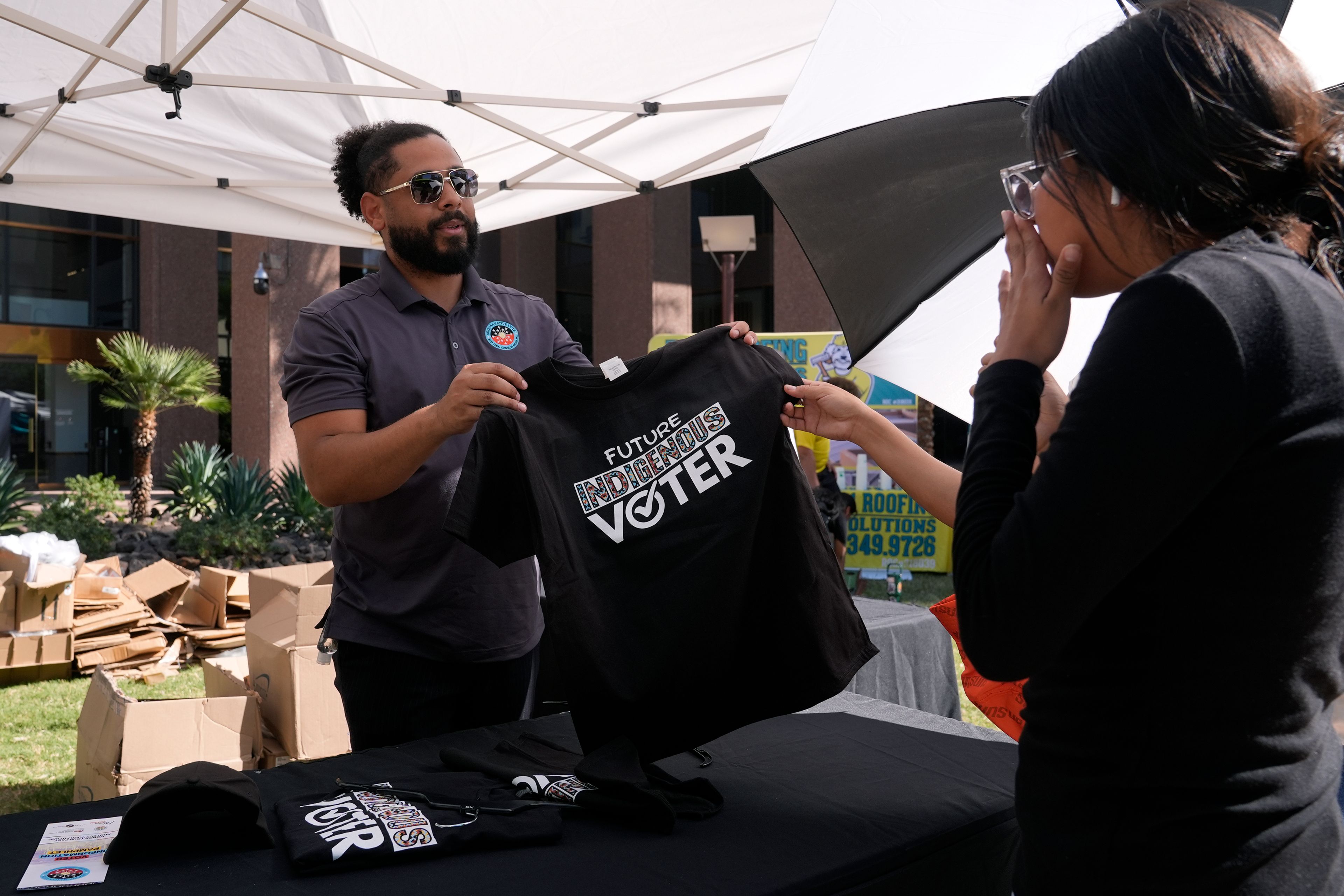 Alexander Castillo-Nunez, left, a civic engagement coordinator at the Inter Tribal Council of Arizona, Inc., shows a voter t-shirt at an Arizona Native Vote booth during an Indigenous Peoples' Day event Monday, Oct. 14, 2024, in Phoenix. (AP Photo/Ross D. Franklin)