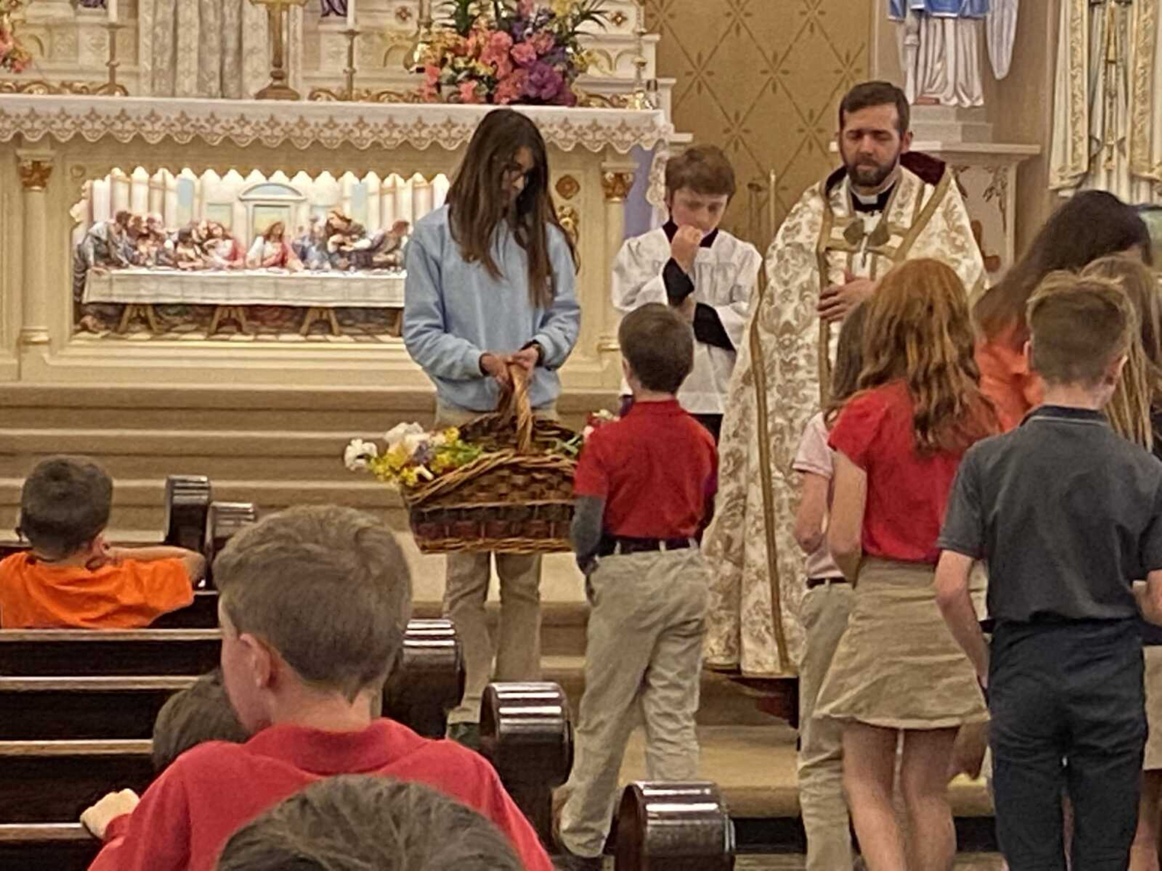 The students and staff of Guardian Angel School process one by one to place each flower in a basket held by Rylie Priggel.  The flowers will be placed in a vase to be put before Mary.  The server is Kasyn Seabaugh who will assist Fr. Kelly with the ceremony.