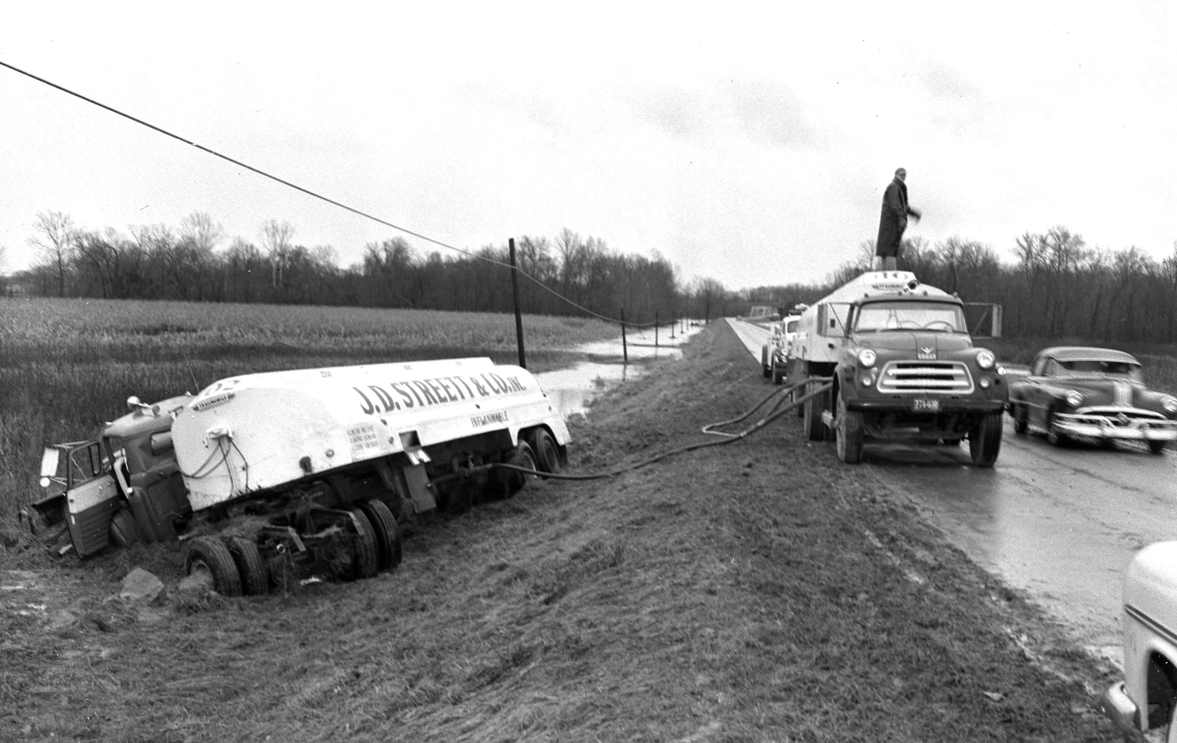 A badly damaged new tractor and trailer owned by the Juden Transportation Co. and hauling 5,200 gallons of gasoline for the J.D. Streett and Co., went off Highway 70 west of the Byrds Creek bridge early Monday, Nov. 18, 1957. The driver, John Kelley, reported that water over the highway obscured his vision when it splashed onto the windshield. A relief truck is shown pumping the fuel from the loaded tanker. In the distant background is the creek bridge.