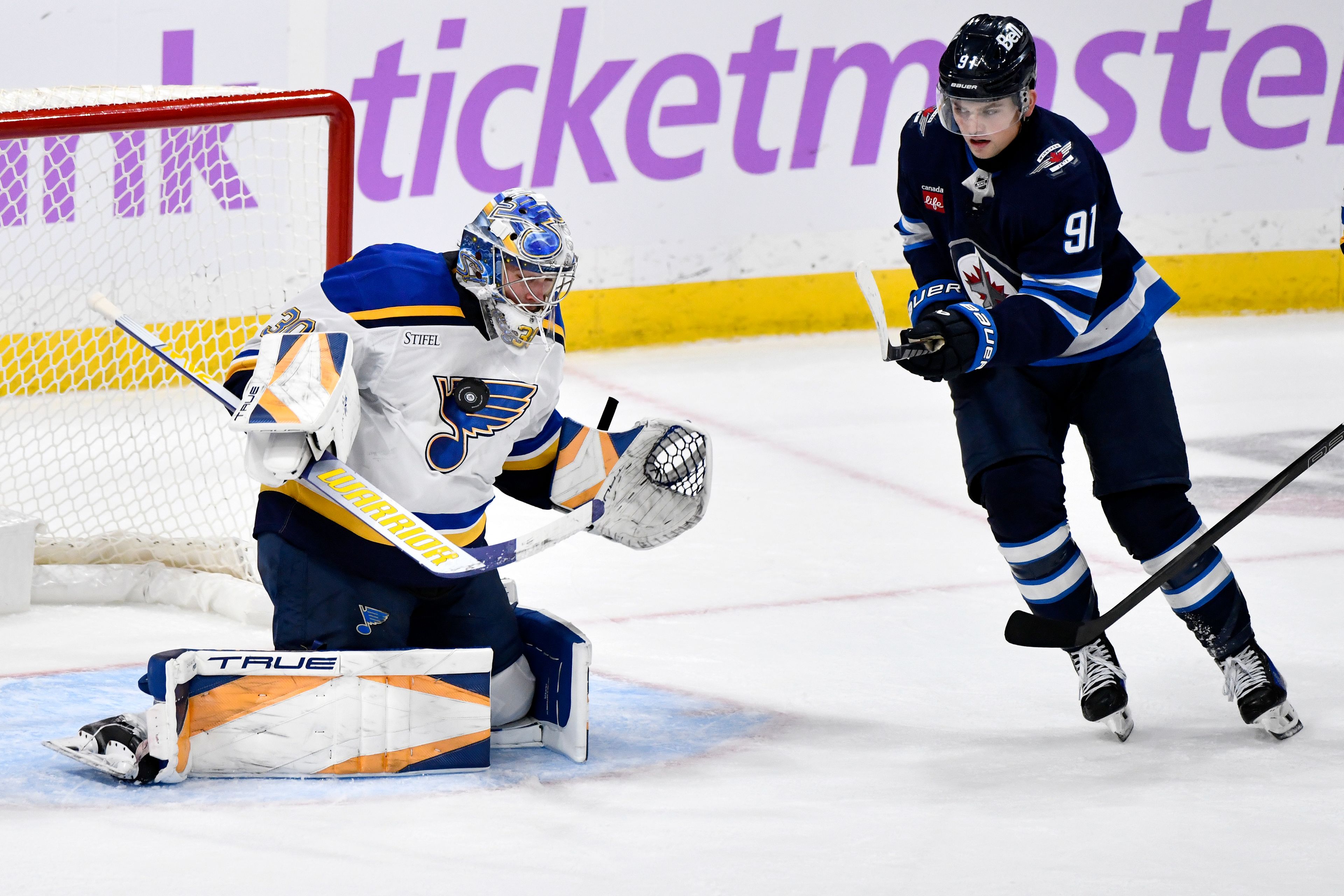 St. Louis Blues goaltender Joel Hofer (30) makes a save on a Winnipeg Jets shot as Cole Perfetti (91) looks for the rebound during the third period of their NHL hockey game in Winnipeg, Canada Tuesday, Dec. 3, 2024. (Fred Greenslade/The Canadian Press via AP)