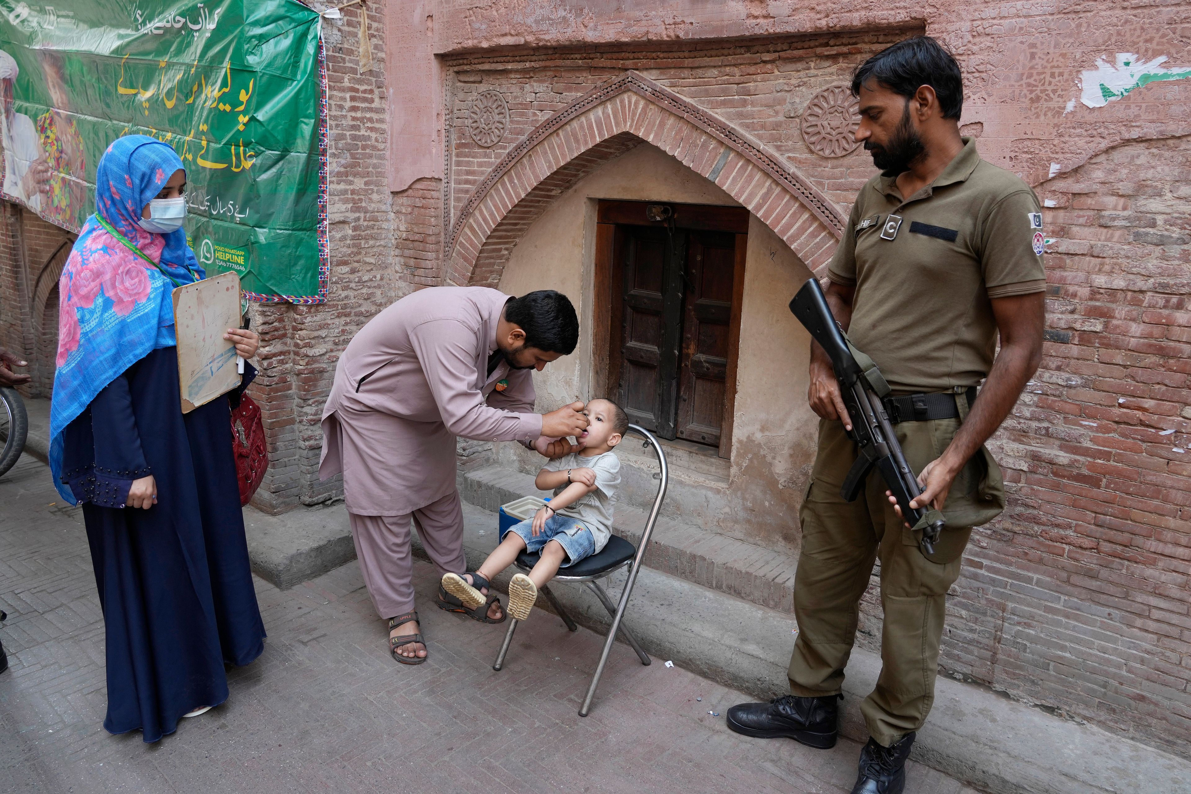 A police officer stands guard as a health worker, center, administers a polio vaccine to a child at a neighbourhood of Lahore, Pakistan, Monday, Oct. 28, 2024. (AP Photo/K.M. Chaudary)