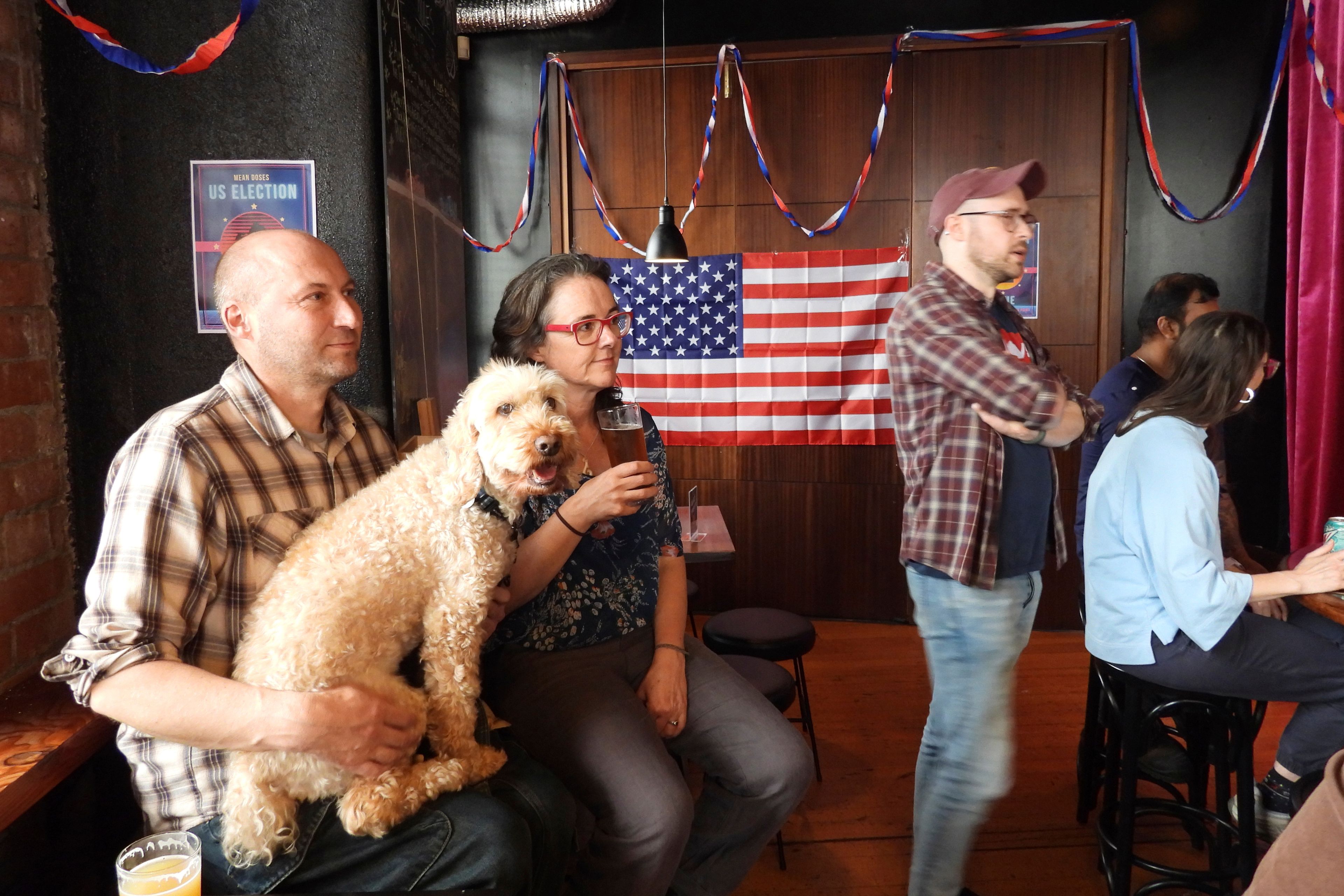 Steve Baker, left, Claudine Earley and their dog Louis watch a television broadcast during a U.S. election viewing party at Mean Doses bar in Wellington, New Zealand on Wednesday, Nov. 6, 2024. (AP Photo/Charlotte Graham-McLay)