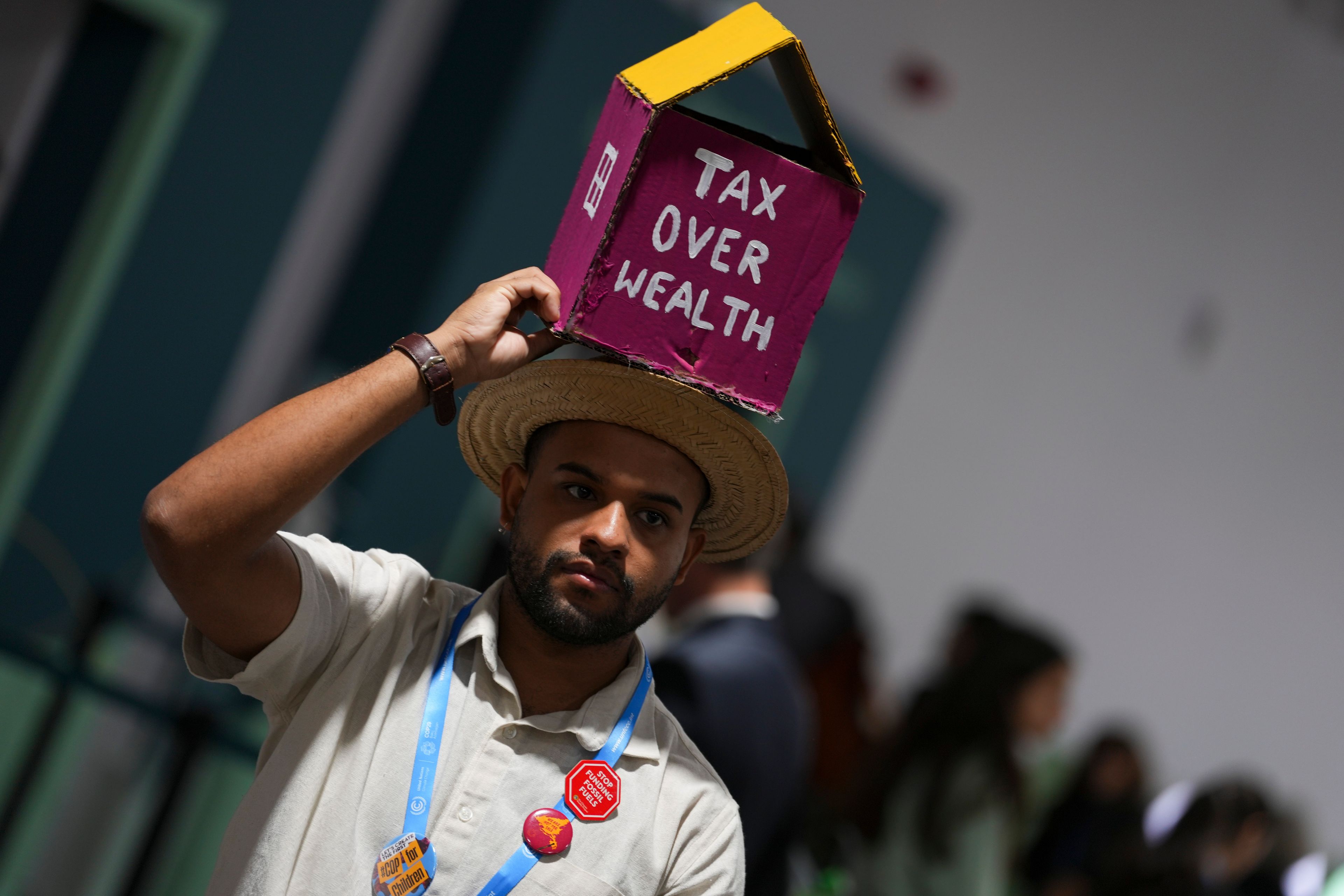 A person walks holding an object with the likeness of a house that says tax over health at the COP29 U.N. Climate Summit, Saturday, Nov. 16, 2024, in Baku, Azerbaijan. (AP Photo/Peter Dejong)