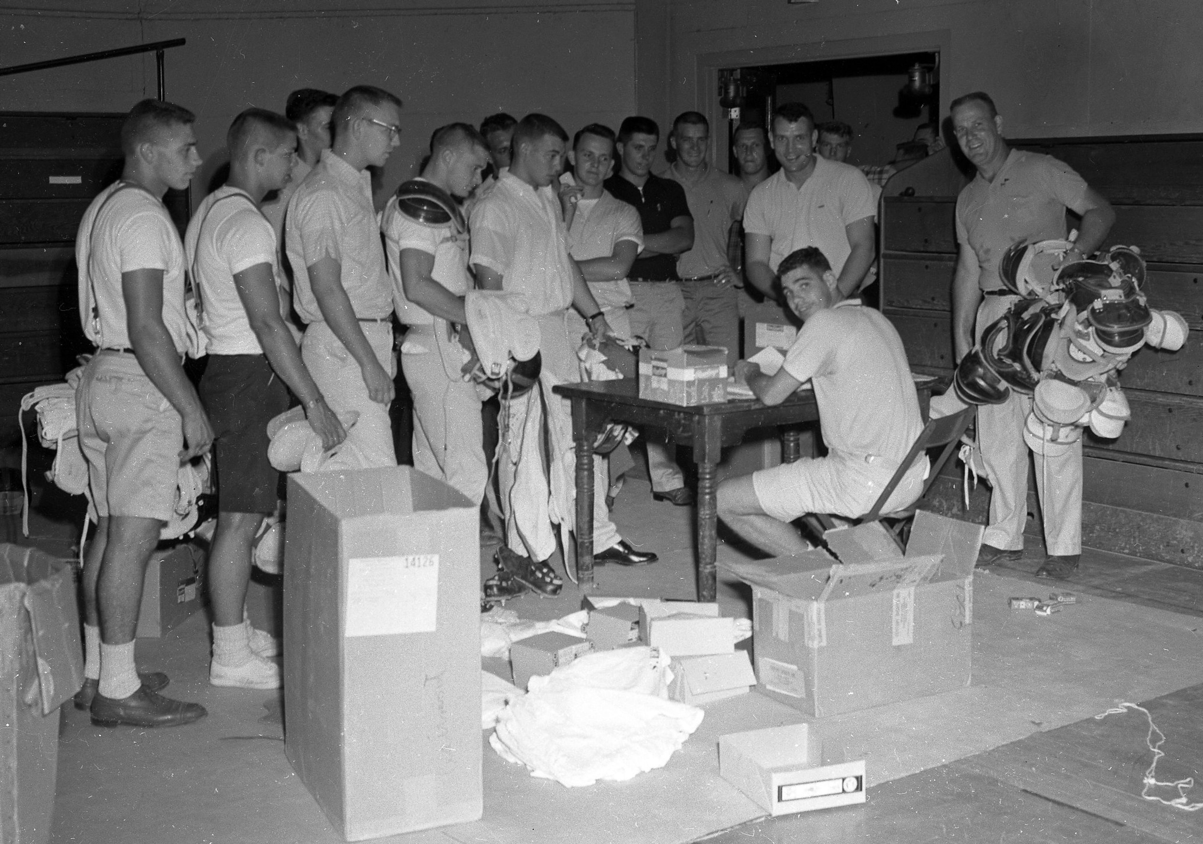 A big crop of State College freshmen, husky ones, too, report Sunday, Aug. 28, 1960, at Houck Field House to receive football equipment for the grid season. At the right, Coach Kenneth Knox holds a bundle of shoulder pads and seated at the desk is assistant coach John Schneider. Standing above Schneider is Jim Hamby, also of the coaching staff.
