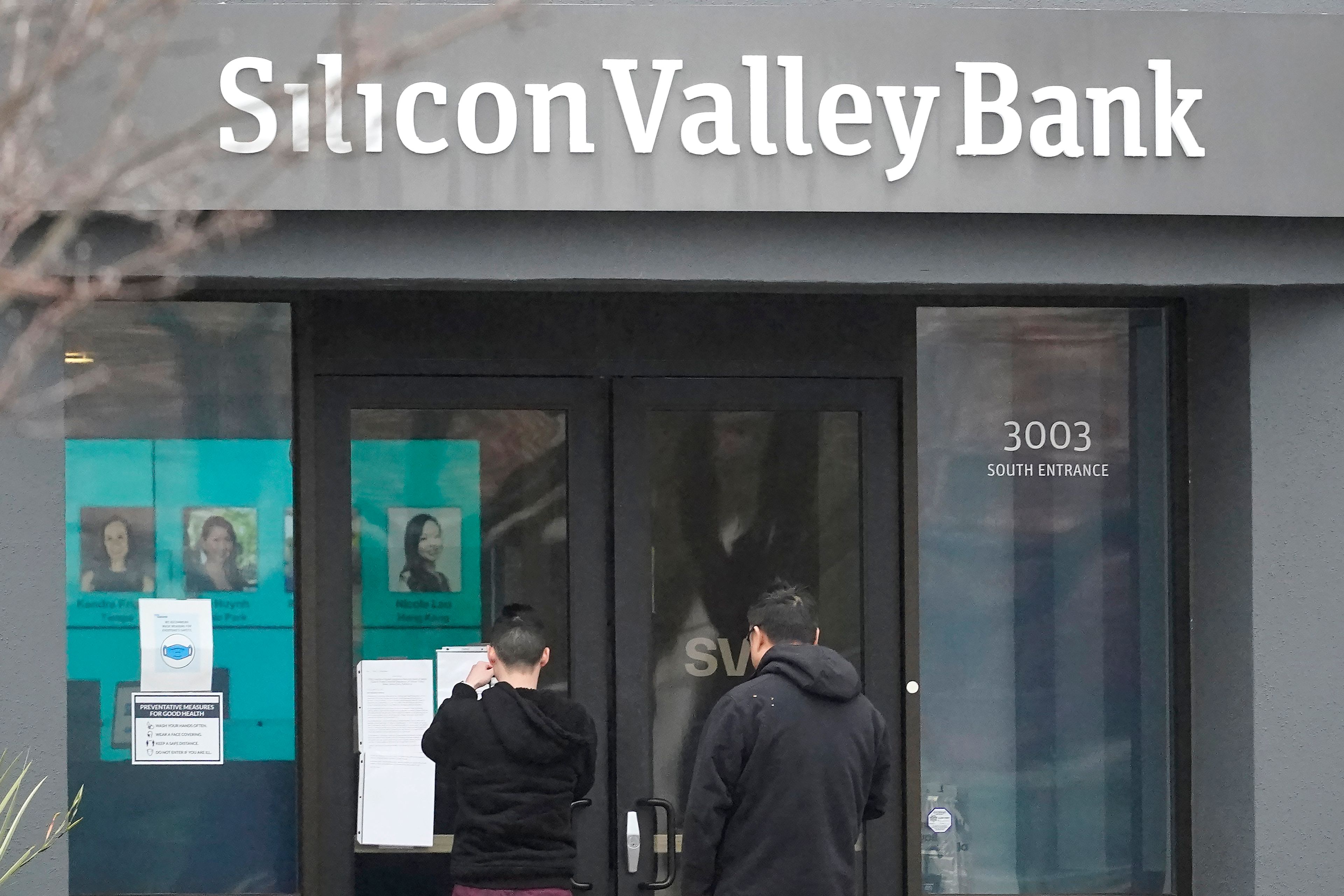 People look at signs posted outside of an entrance to Silicon Valley Bank in Santa Clara, Calif., Friday, March 10, 2023. The Federal Deposit Insurance Corporation is seizing the assets of Silicon Valley Bank, marking the largest bank failure since Washington Mutual during the height of the 2008 financial crisis. The FDIC ordered the closure of Silicon Valley Bank and immediately took position of all deposits at the bank Friday. (AP Photo/Jeff Chiu)