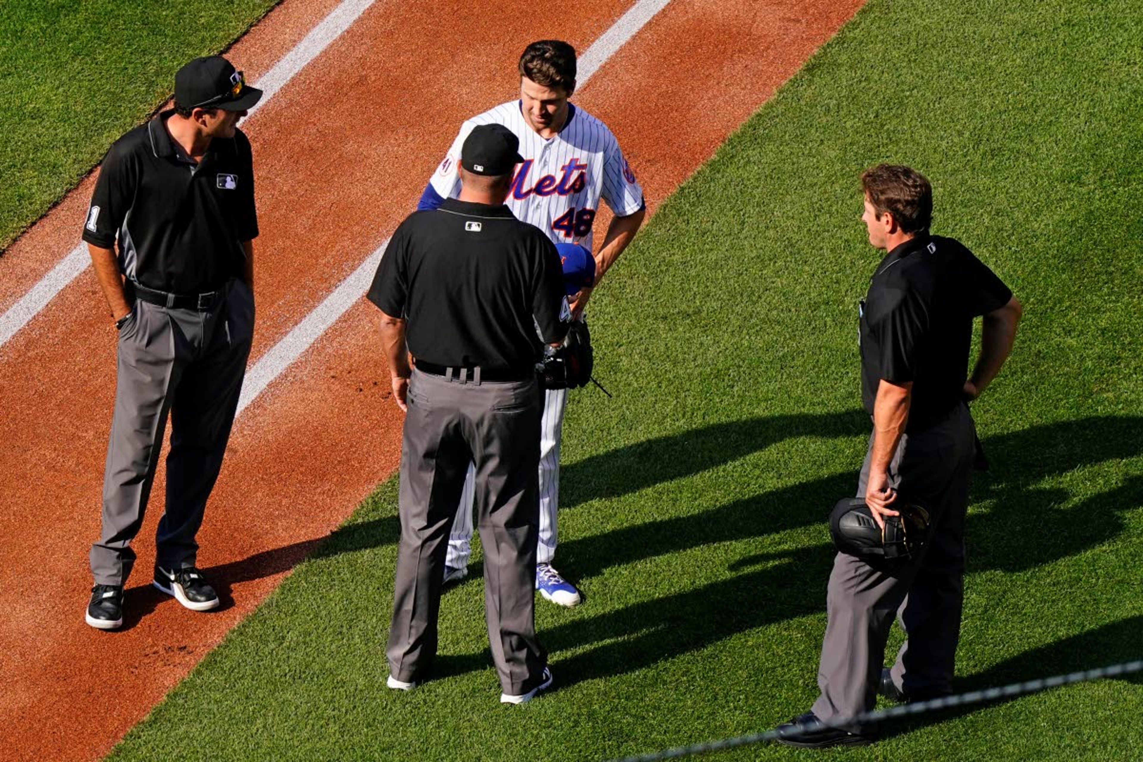 Third base umpire Ron Kulpa, bottom center, asks New York Mets starter Jacob deGrom, top center, about foreign substances before deGrom headed to the dugout after pitching in the first inning of a baseball game against the Atlanta Braves, Monday, June 21, 2021, in New York. Home plate umpire Ben May, right, looks on. (AP Photo/Kathy Willens)