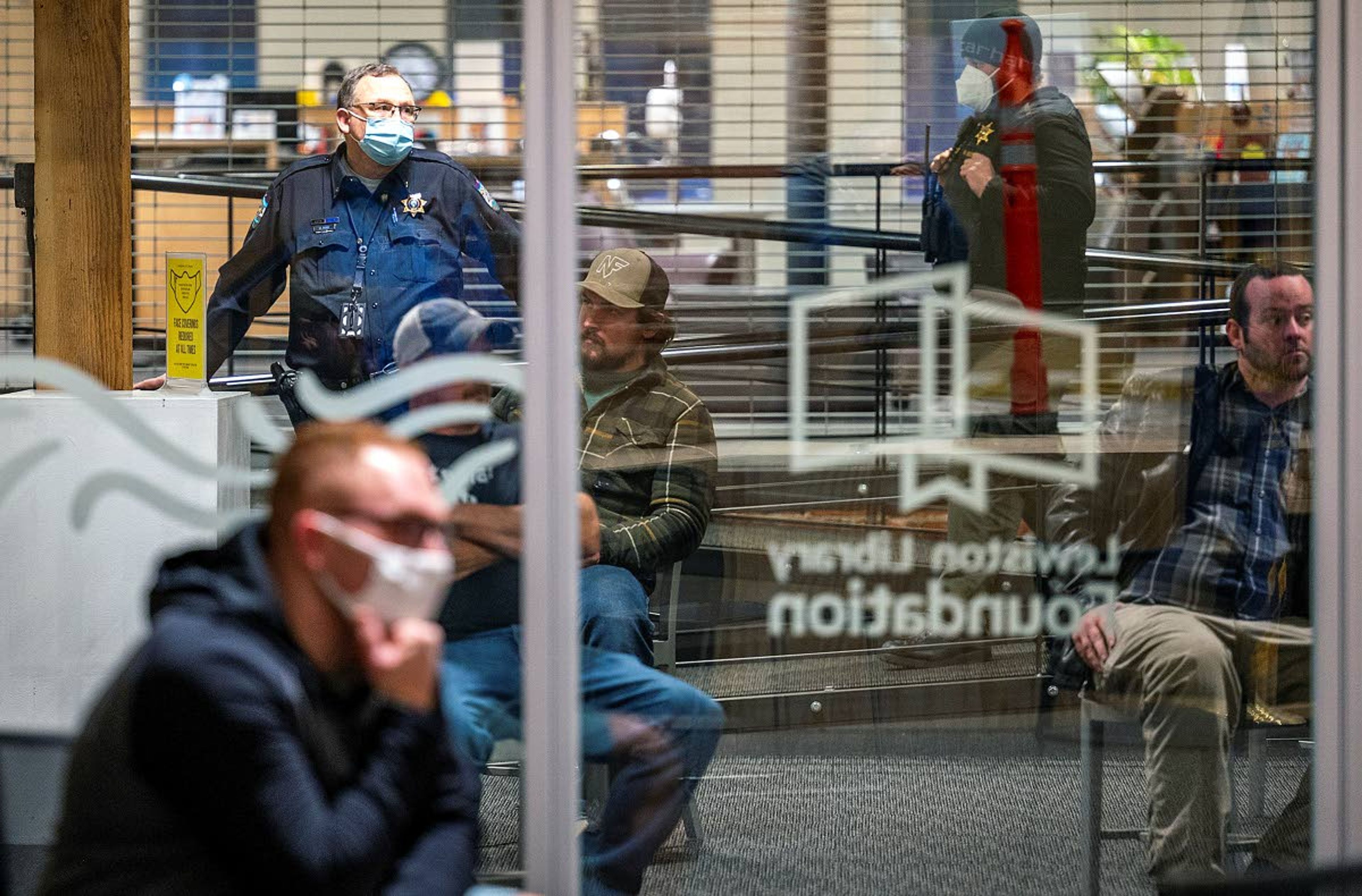 A few people wear masks and others refrain from wearing masks as Lewiston Police Chief Budd Hurd (left) watches from outside the meeting room as people wait their turn to enter and make a public comment during a Lewiston City Council special meeting on Thursday at the Lewiston City Library.