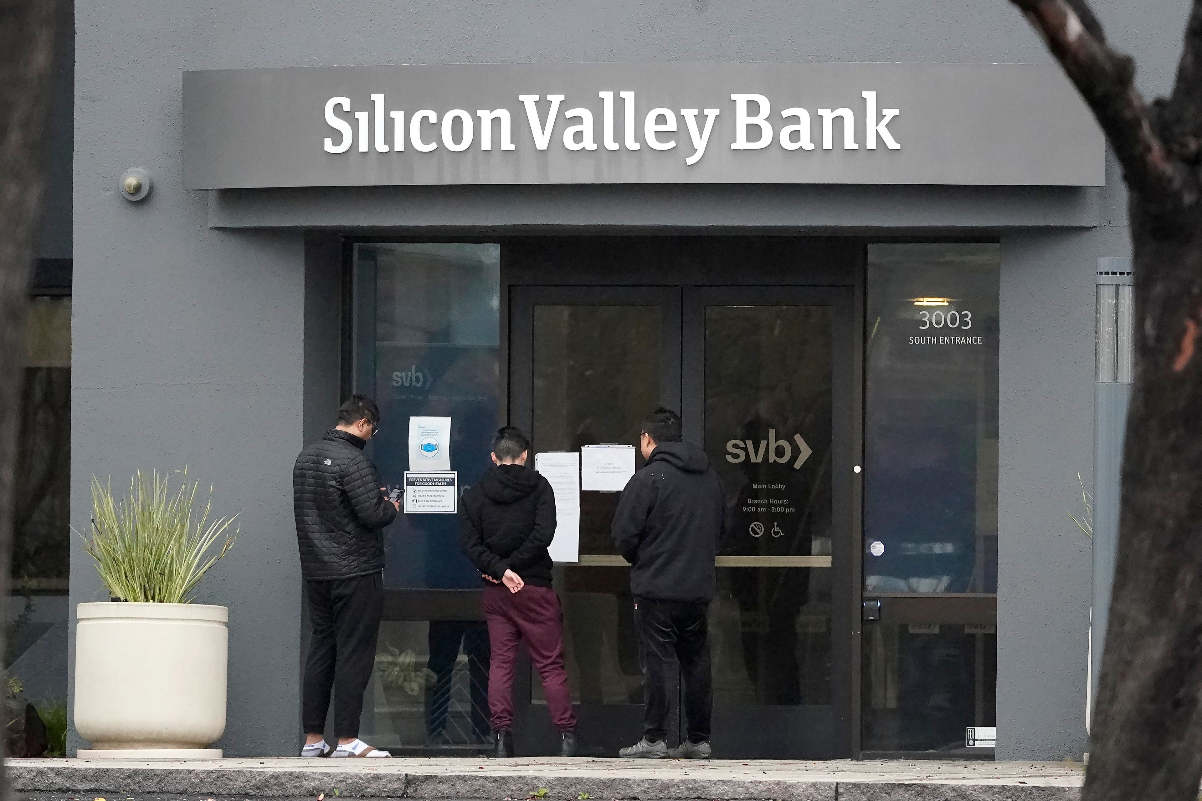 People look at signs posted outside of an entrance to Silicon Valley Bank in Santa Clara, Calif., Friday, March 10, 2023. The Federal Deposit Insurance Corporation is seizing the assets of Silicon Valley Bank, marking the largest bank failure since Washington Mutual during the height of the 2008 financial crisis. The FDIC ordered the closure of Silicon Valley Bank and immediately took position of all deposits at the bank Friday. (AP Photo/Jeff Chiu)