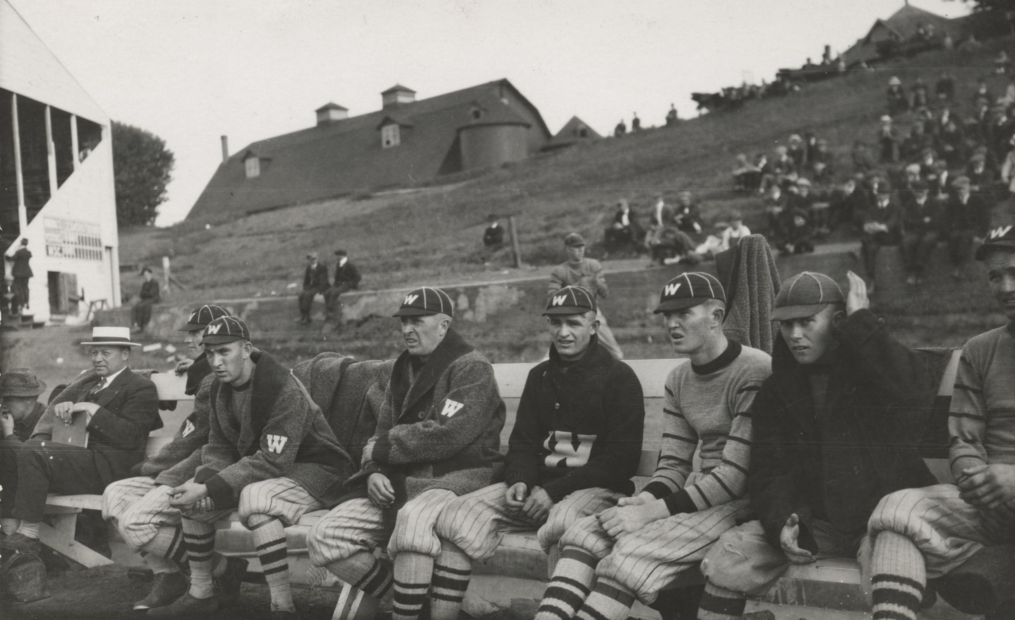 Washington State College players watch the action from the bench, as fans sit on the hillside in the background. This location is the current south stands of Martin Stadium.