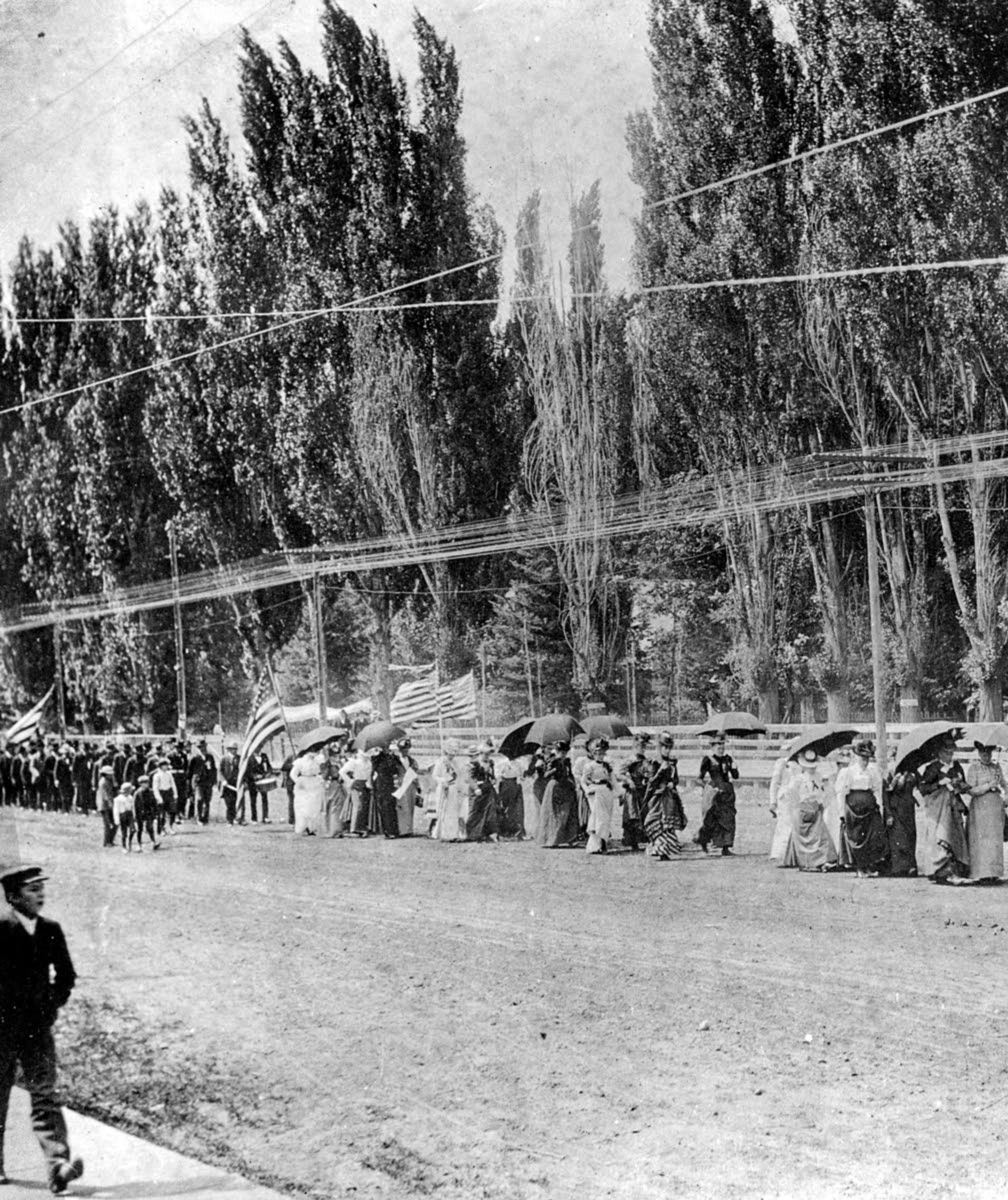 In this July 1896 photo, a line of women march on a dirt road, holding flags and umbrellas for a suffrage parade in Lewiston. Following them is a line of men and boys.