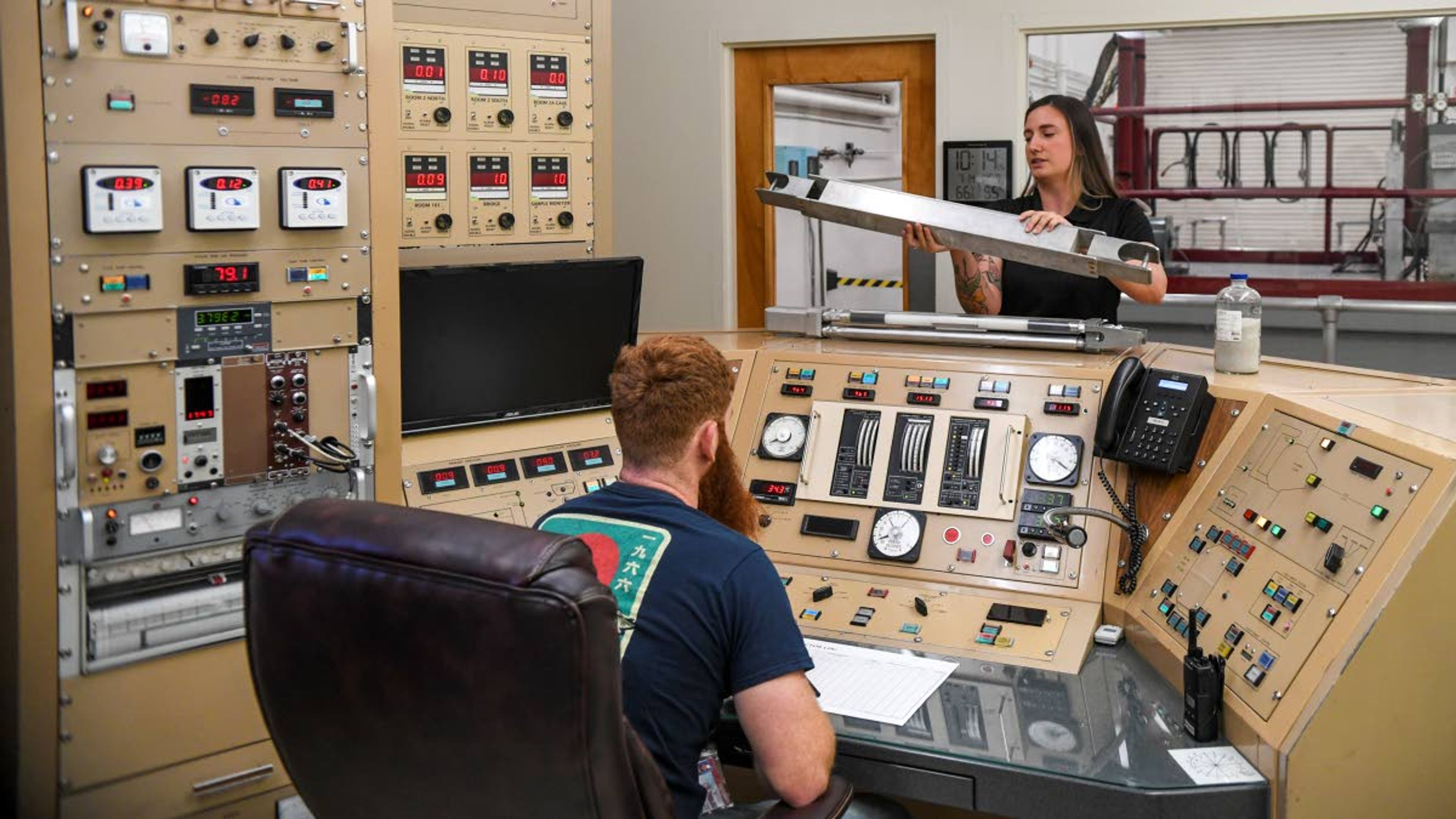 Research operations engineer Maddison Heine, back, holds a mock-up of the original plate-type fuel design used in the nuclear reactor while trainee Derrick Adams watches the control panel inside the reactor control room at Washington State University’s Nuclear Science Center.