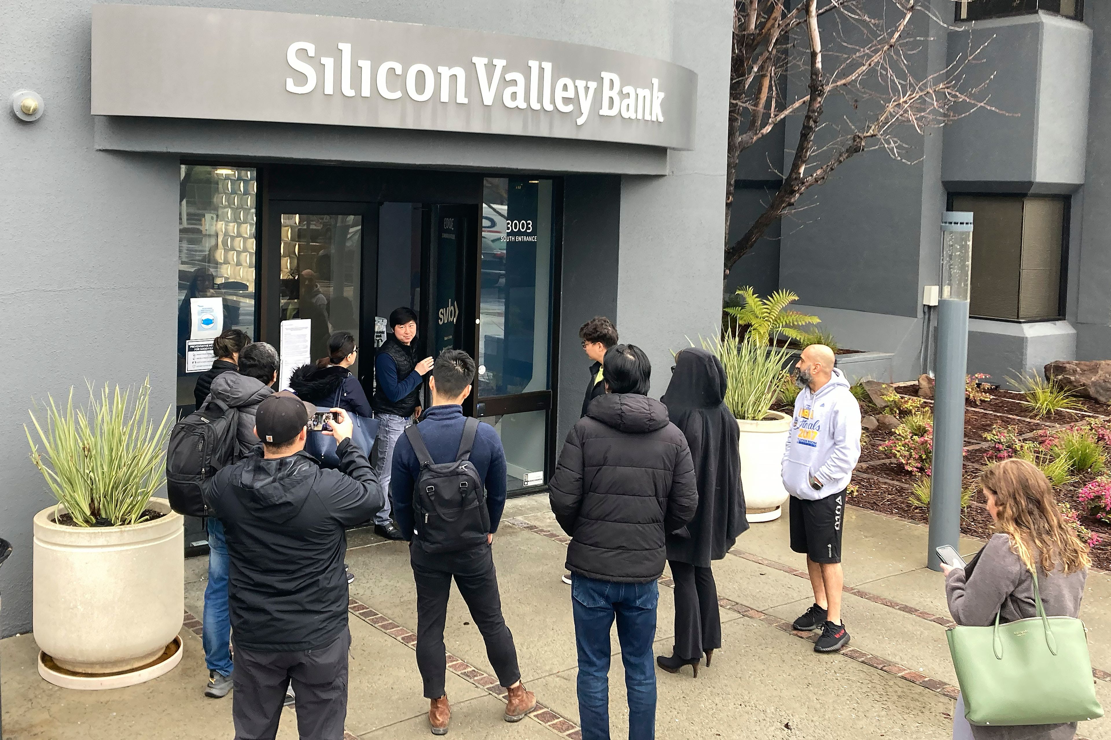 A person from inside Silicon Valley Bank, middle rear, talks to people waiting outside of an entrance to Silicon Valley Bank in Santa Clara, Calif., Friday, March 10, 2023. The Federal Deposit Insurance Corporation seized the assets of the bank on Friday, marking the largest bank failure since Washington Mutual during the height of the 2008 financial crisis. (AP Photo/Jeff Chiu)