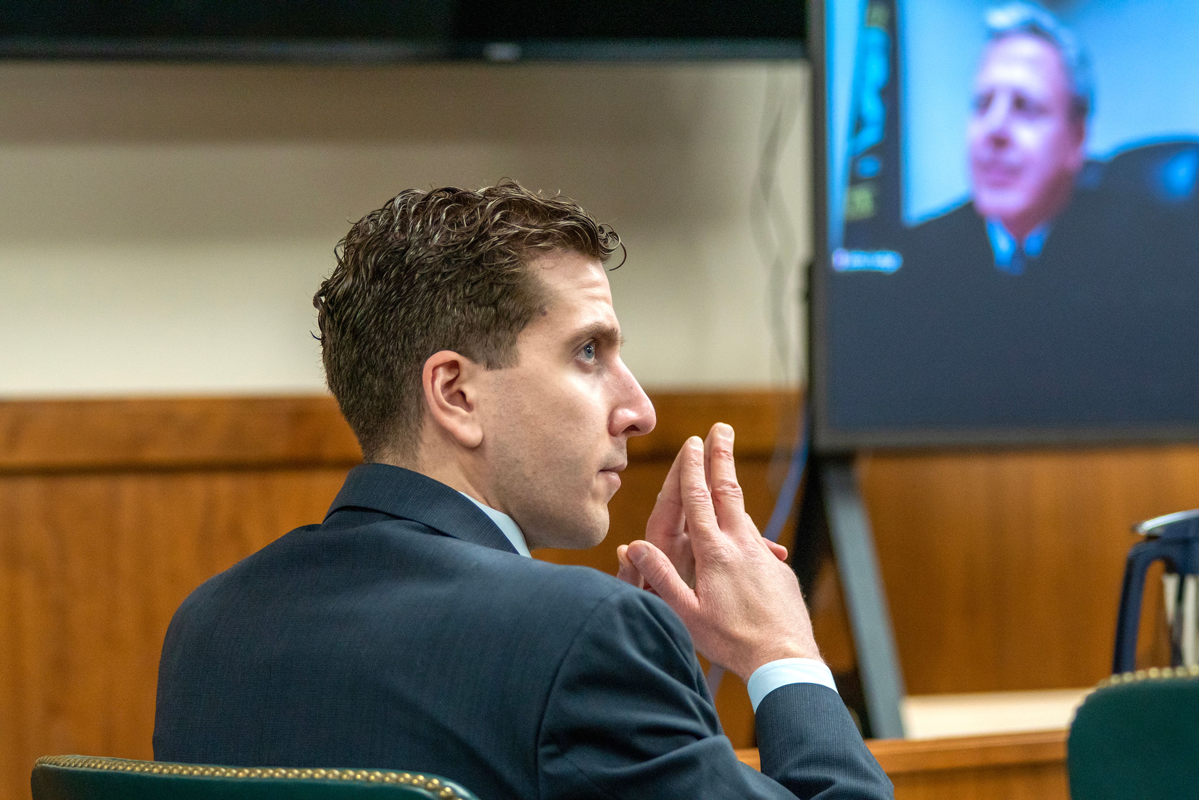 Quadruple homicide suspect Bryan Kohberger listens to arguments during a hearing in October of 2023, with Judge John Judge's image appearing on a television screen.