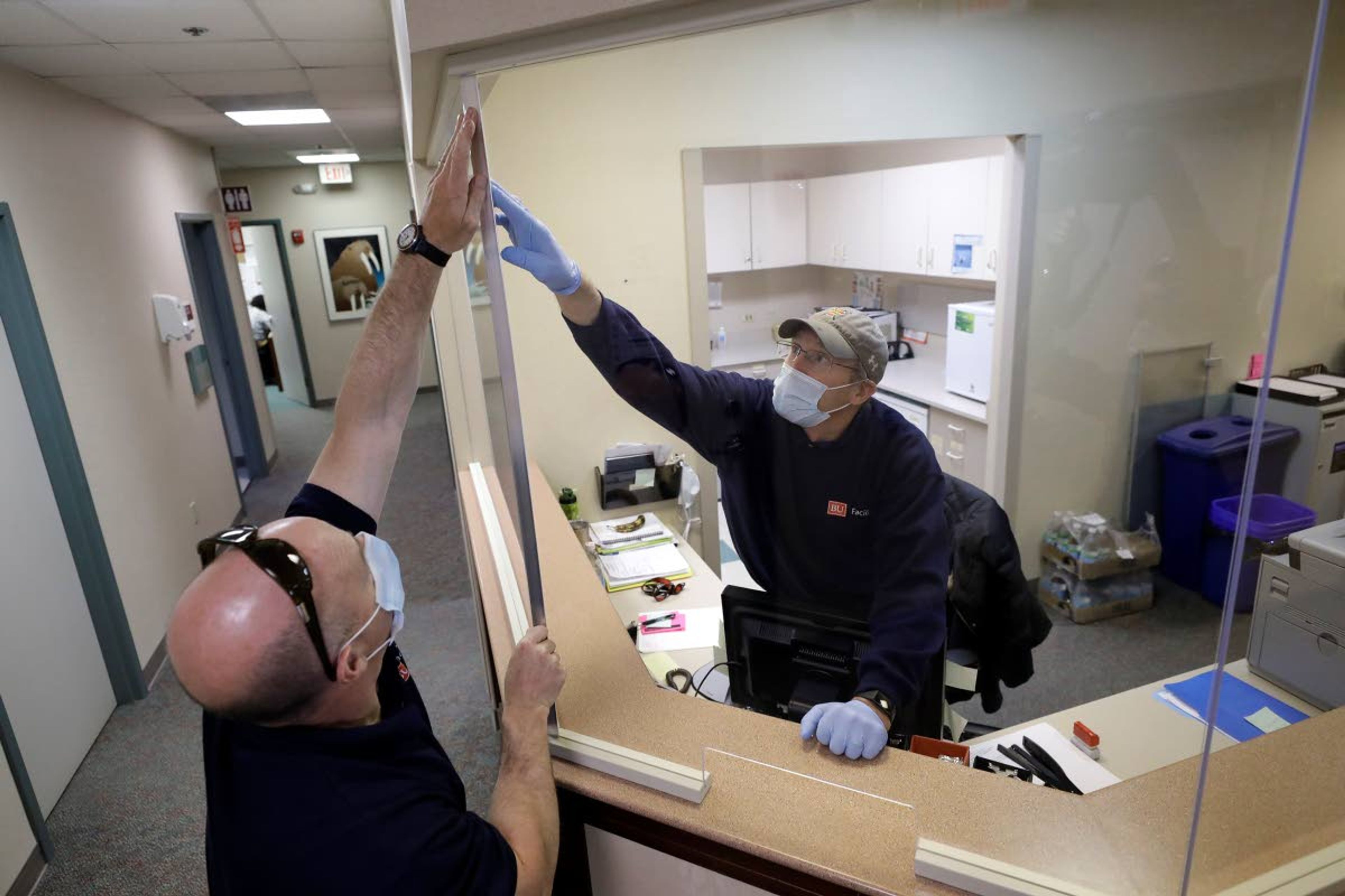 In this Wednesday, May 20, 2020 photo carpenters John Mackie, of Canton, Mass., left, and Doug Hathaway, of Holliston, Mass., right, apply trim to a newly installed plastic barrier in an office area, at Boston University, in Boston. Boston University is among a growing number of universities making plans to bring students back to campus this fall, but with new measures meant to keep the coronavirus at bay. (AP Photo/Steven Senne)