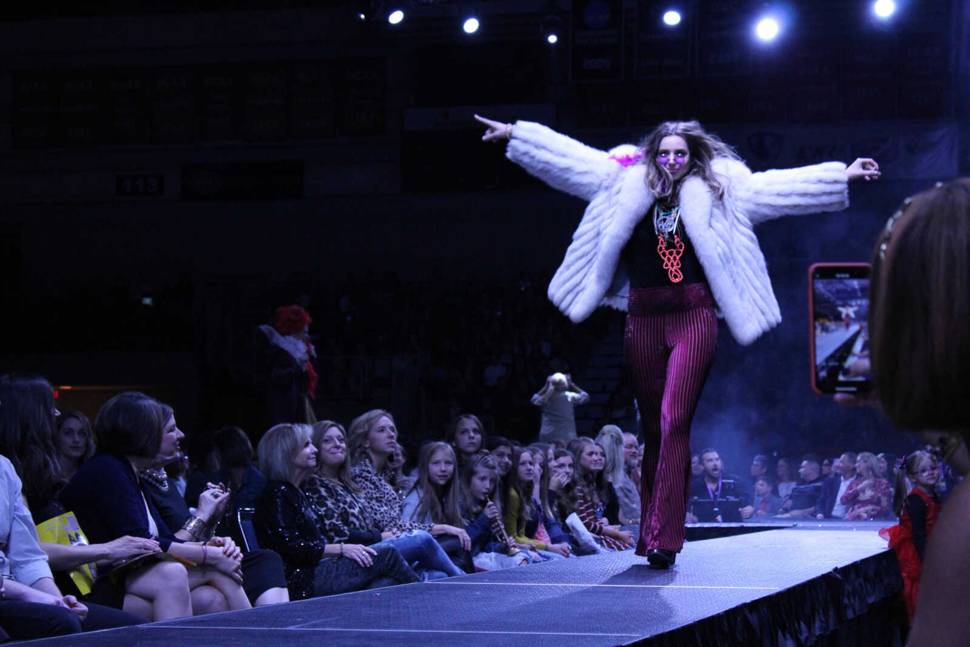 Katelyn Lollas, wearing fuchsia tinted sunglasses, a large fur coat and pleated flare pants, is seen walking the runway as Janis Joplin during the 2019 VintageNOW fashion show.
